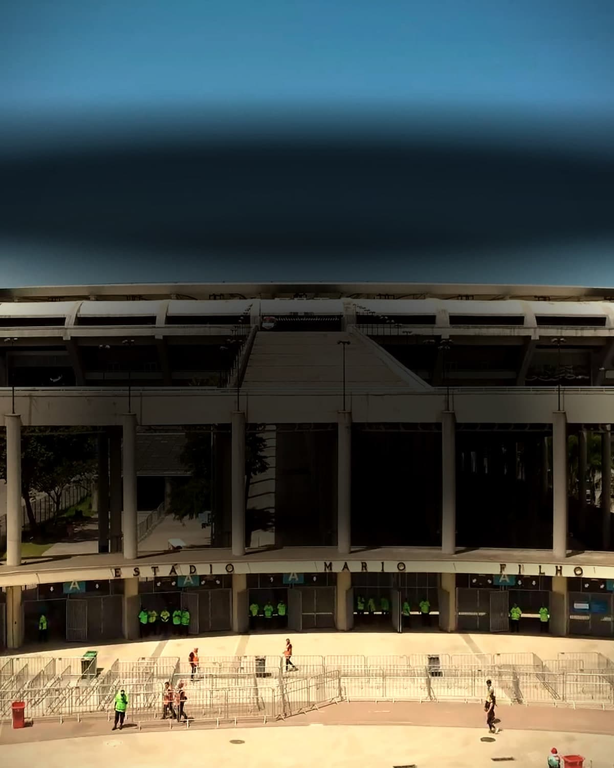 A fachada do Maracanã em dia claro, com a rampa de acesso ao centro, as colunas de concreto, palmeiras e algumas pessoas pequenas na praça em frente.