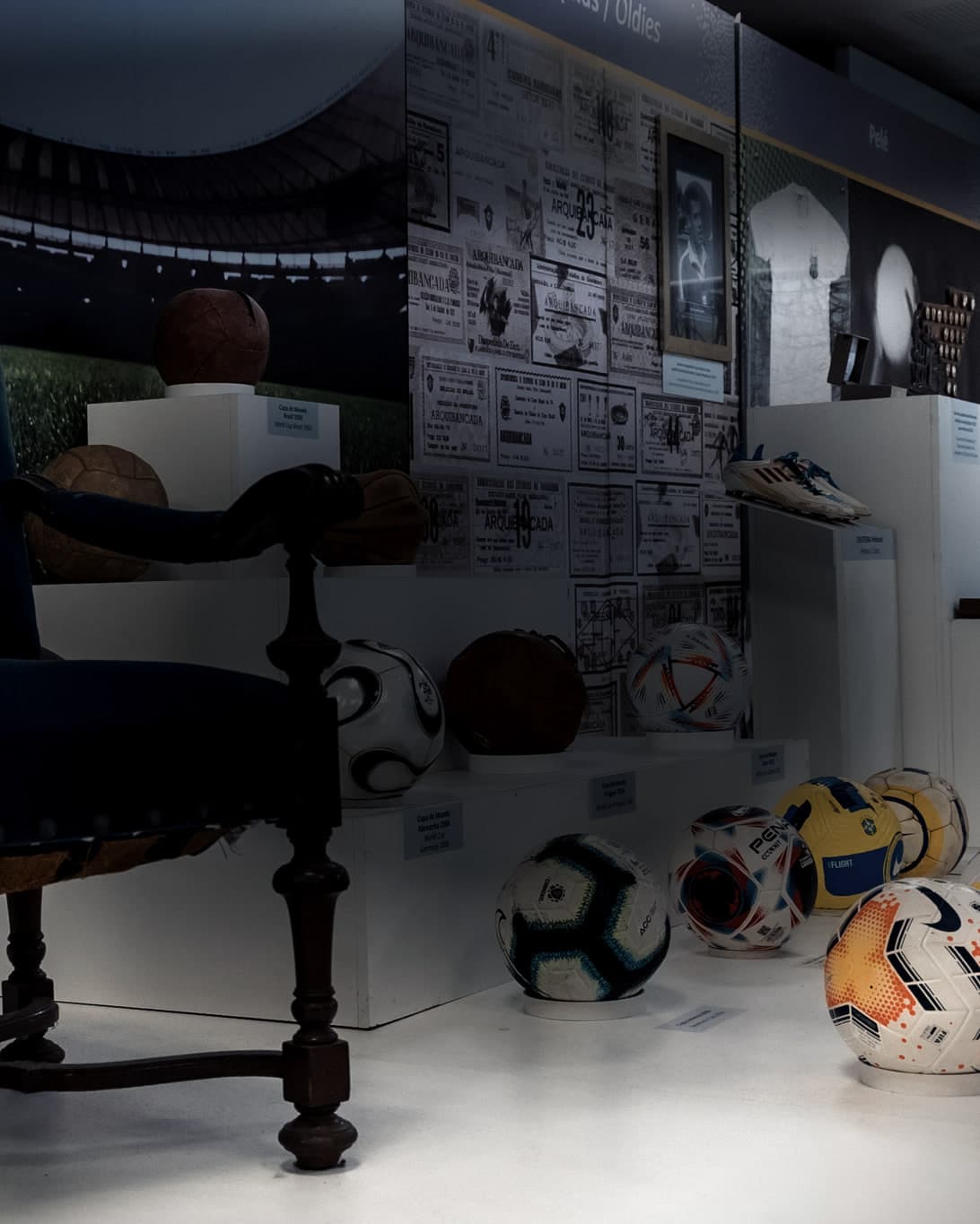 Uma poltrona antiga de veludo azul ao lado de bolas de futebol de várias épocas expostas sobre pedestais brancos, com um painel de ingressos antigos ao fundo.