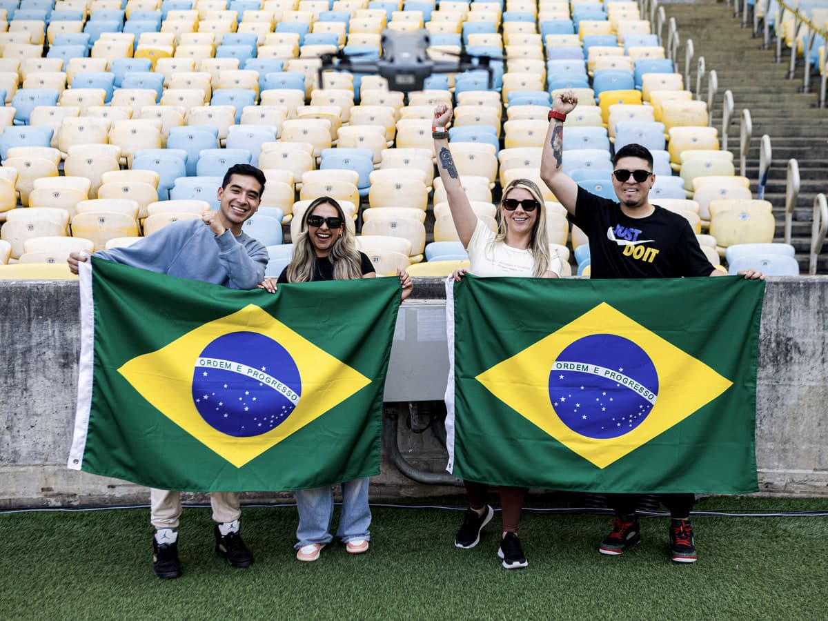 Quatro visitantes na beira do gramado do Maracanã, abrindo duas bandeiras do Brasil diante das cadeiras azuis, amarelas e brancas da arquibancada.