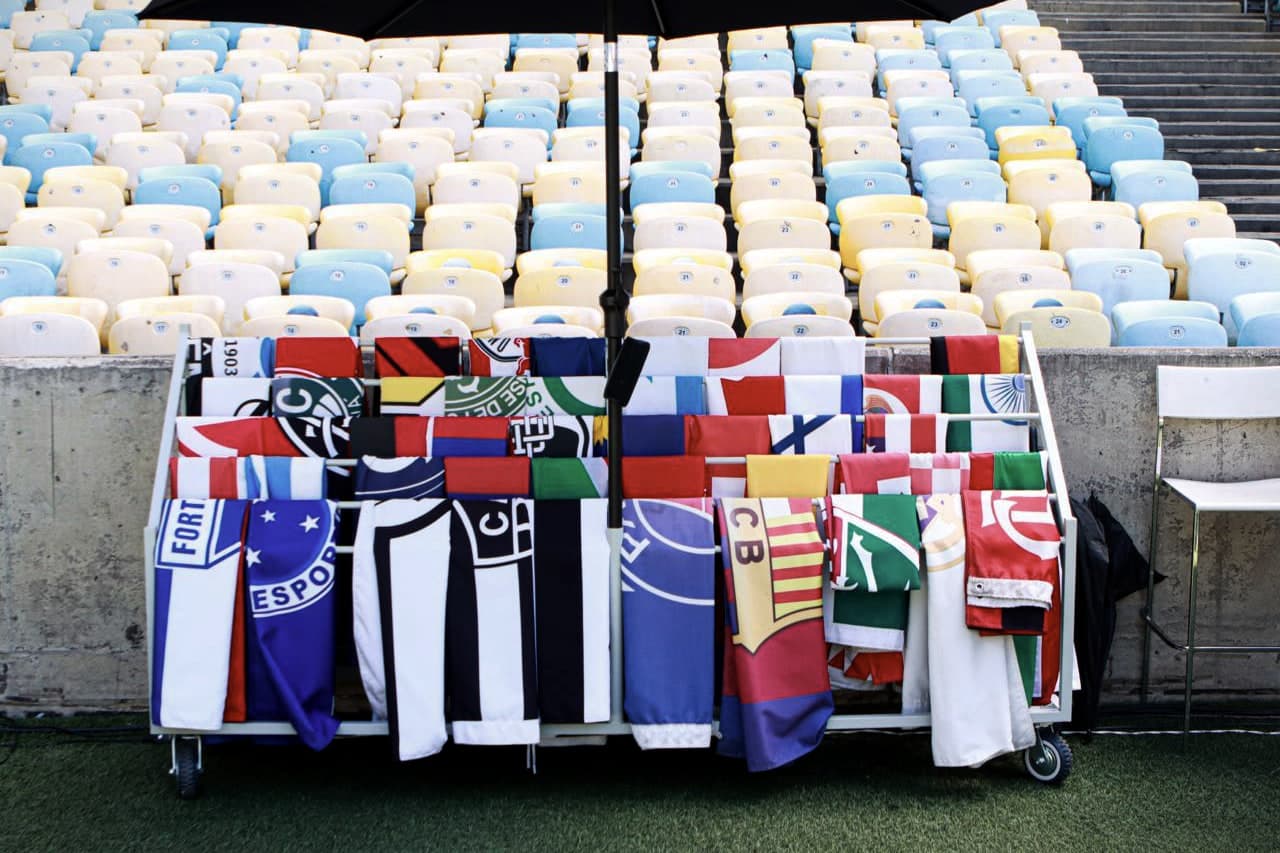 Um carrinho de bandeiras de clubes e países na beira do gramado, sob um guarda-sol, diante das cadeiras coloridas da arquibancada.