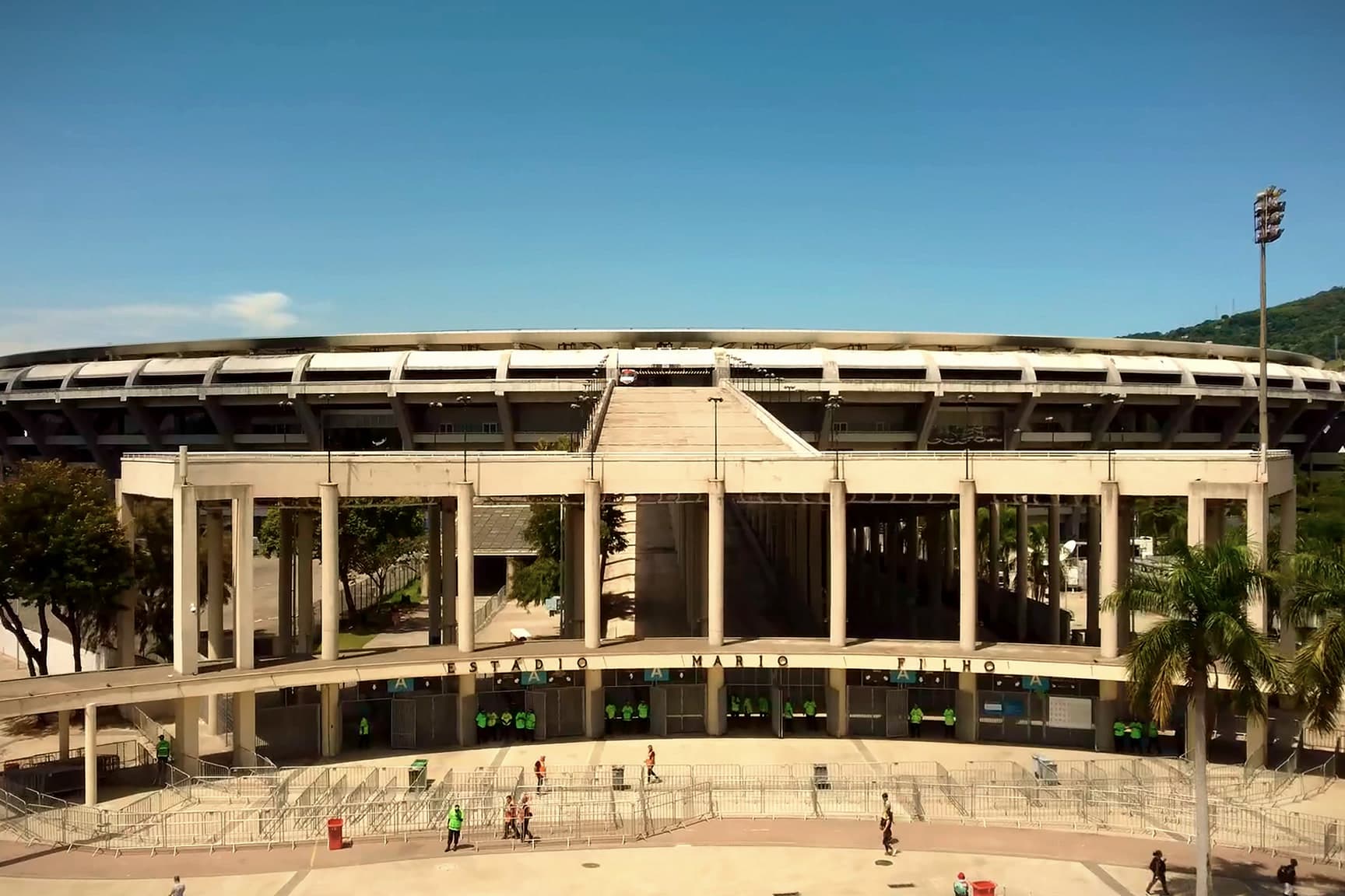 A fachada do Maracanã vista do alto, com a rampa de acesso, as colunas e o letreiro "Estádio Mário Filho", sob o céu azul.