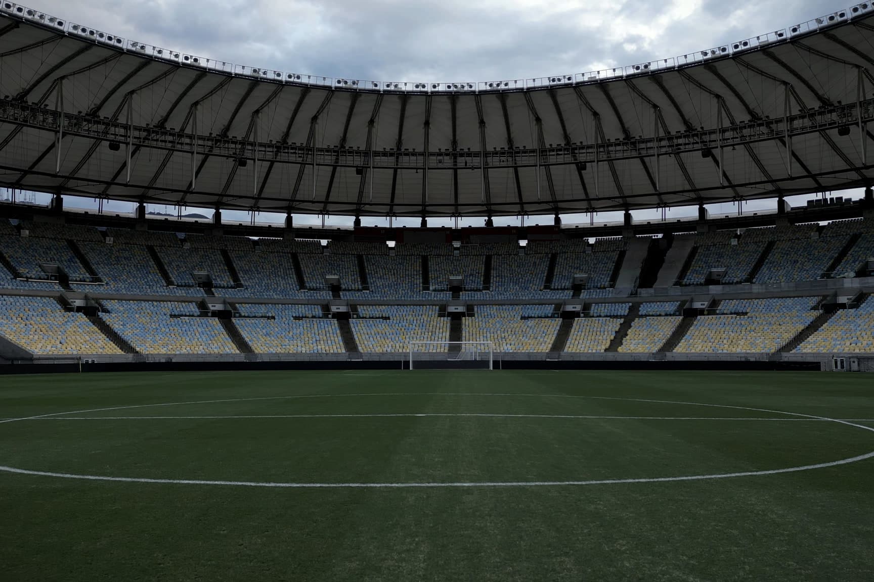 O gramado do Maracanã visto rente à grama, com o círculo central em primeiro plano, o gol ao fundo e o anel de arquibancadas sob a cobertura.