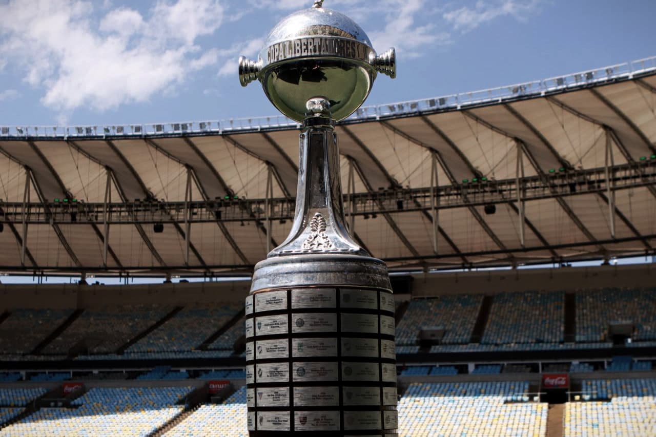 A taça da Libertadores no gramado do Maracanã, com a cobertura e a arquibancada ao fundo sob o céu azul.