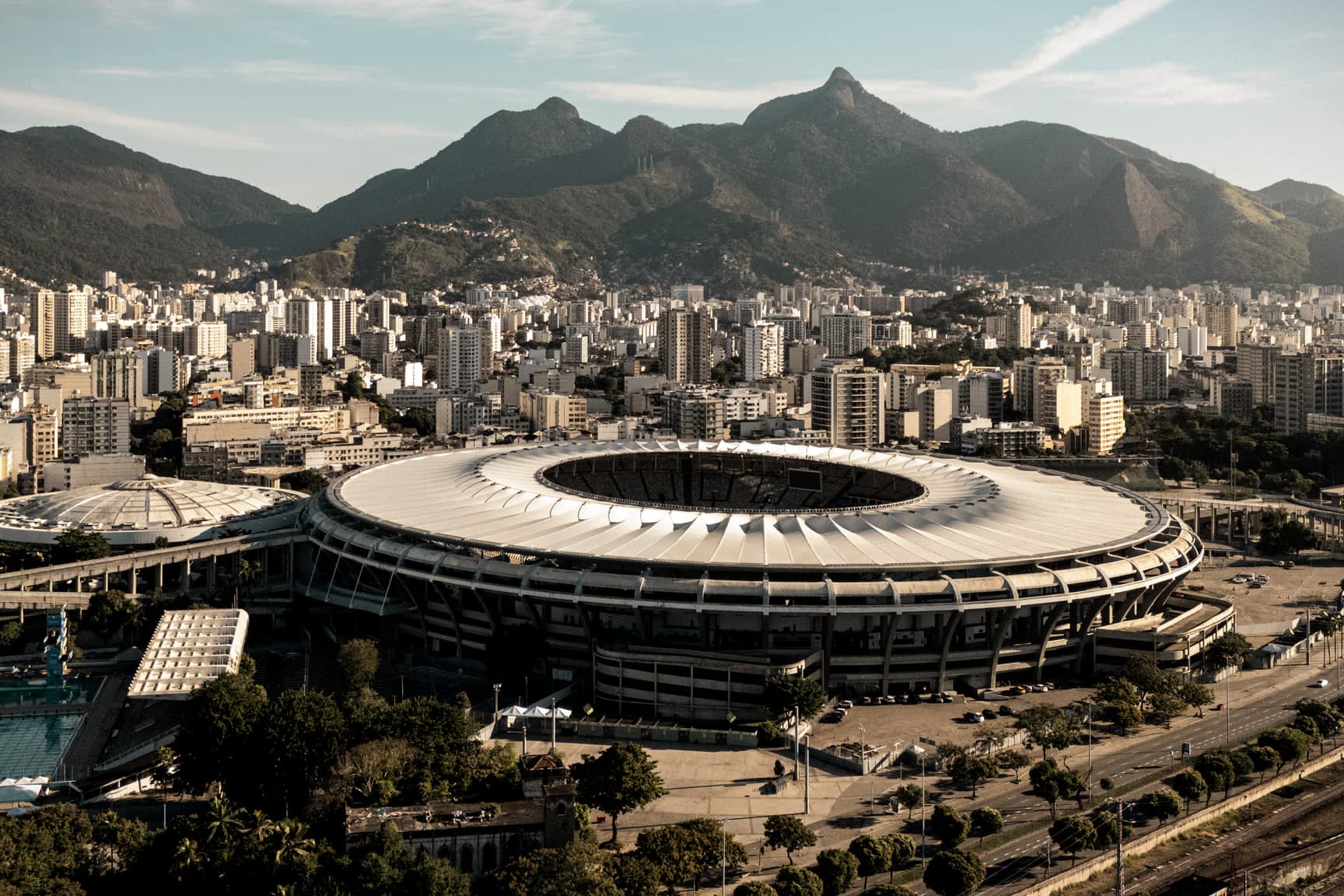 O Maracanã visto do alto em fim de tarde, com o Maracanãzinho ao lado, os prédios do bairro atrás e a serra ao fundo.