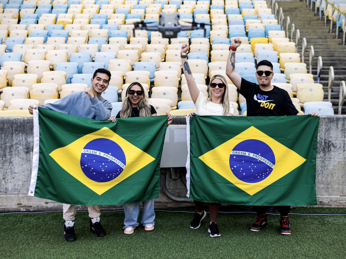 Quatro visitantes na beira do gramado do Maracanã, abrindo duas bandeiras do Brasil diante das cadeiras azuis, amarelas e brancas da arquibancada.