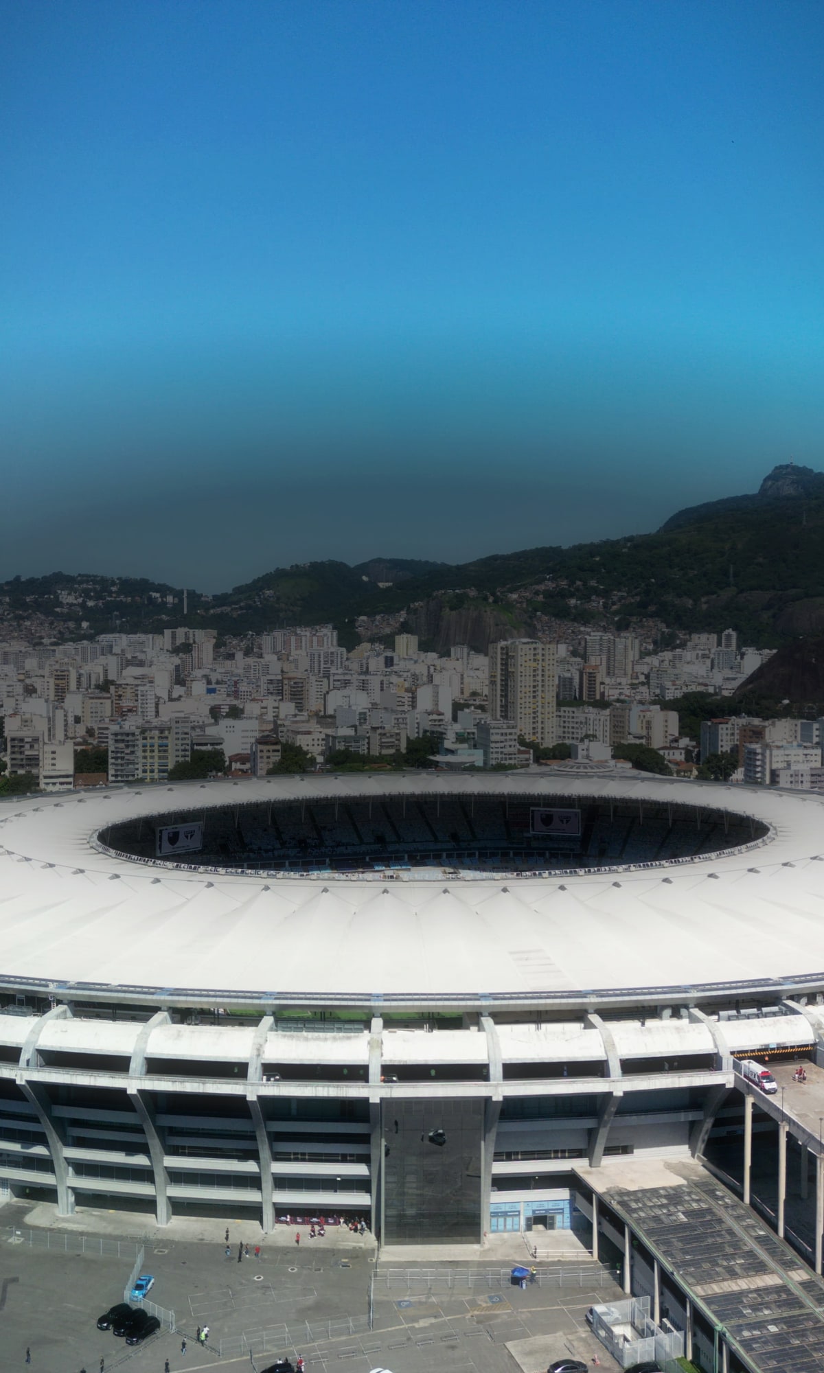 O Maracanã visto do alto em dia claro, a cobertura branca em anel no centro do quadro, os prédios do Rio de Janeiro em volta e a serra da Tijuca ao fundo, sob o céu azul.