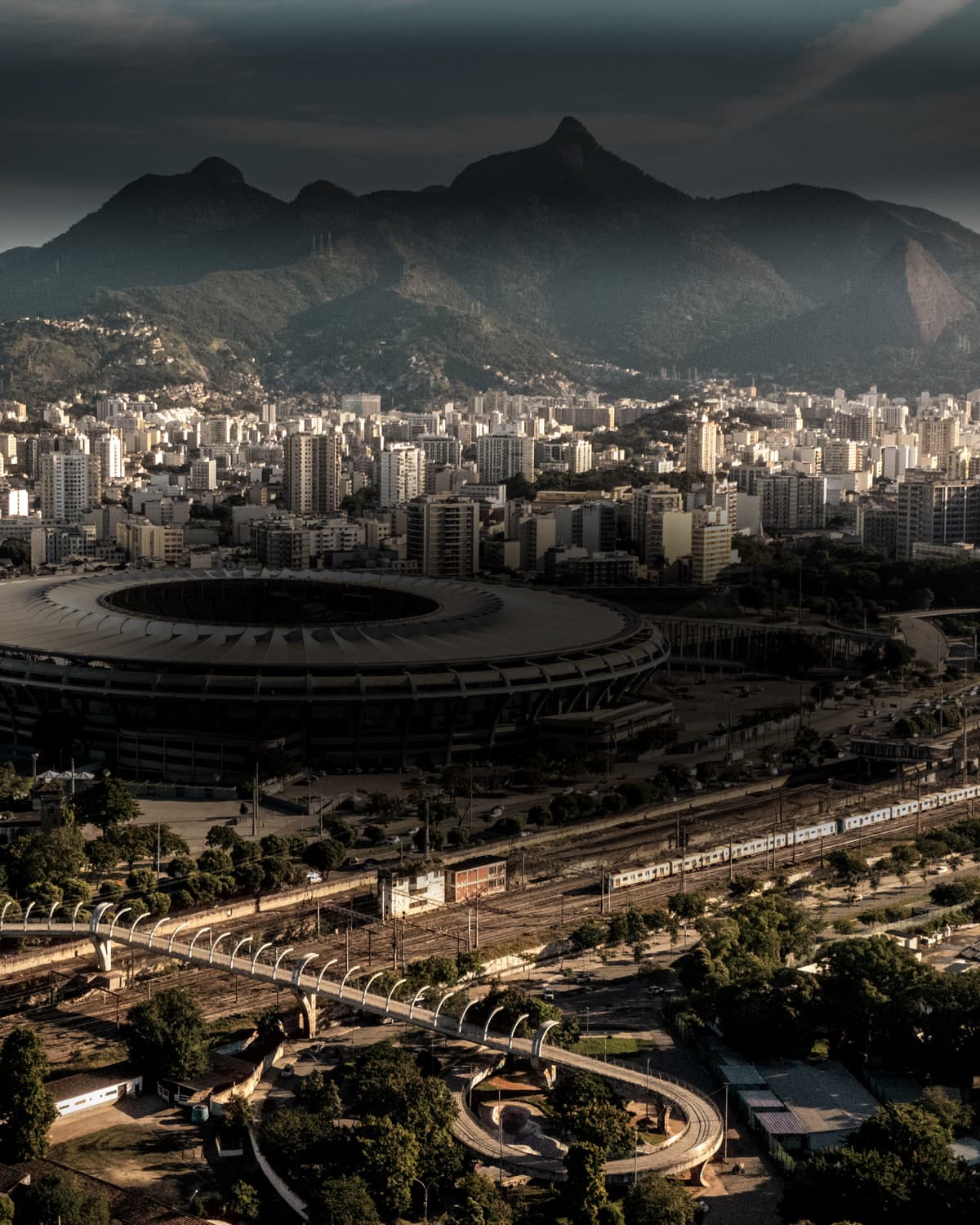 O Maracanã visto do alto no fim da tarde, com a cobertura em anel no terço direito, a estação e as ruas do bairro ao redor e a serra ao fundo.