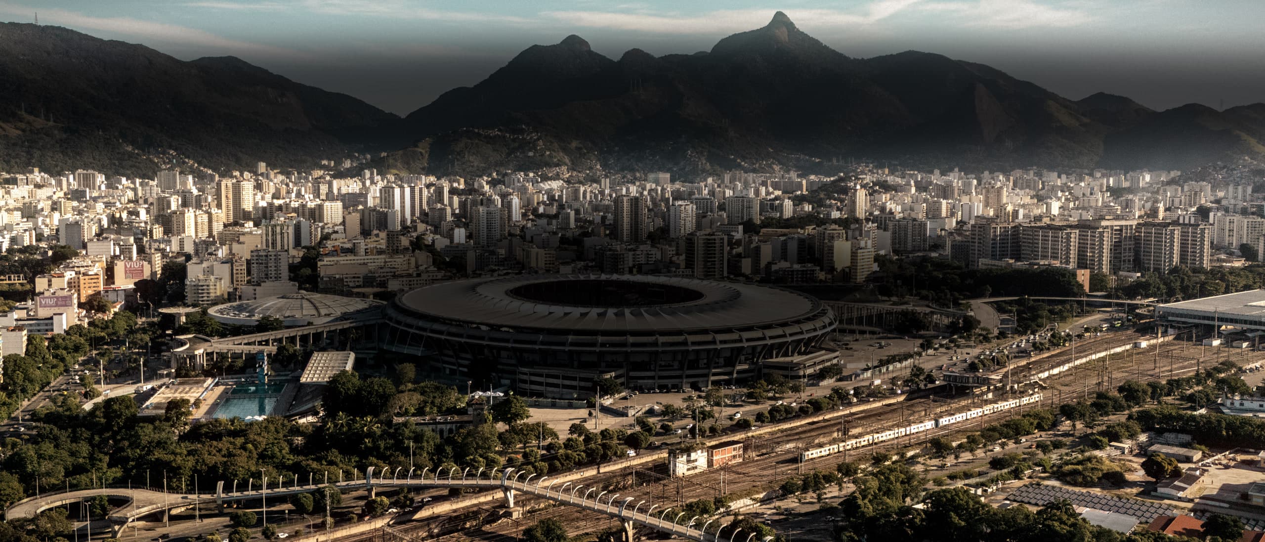 O Maracanã visto do alto no fim da tarde, com a cobertura em anel no terço direito, a estação e as ruas do bairro ao redor e a serra ao fundo.