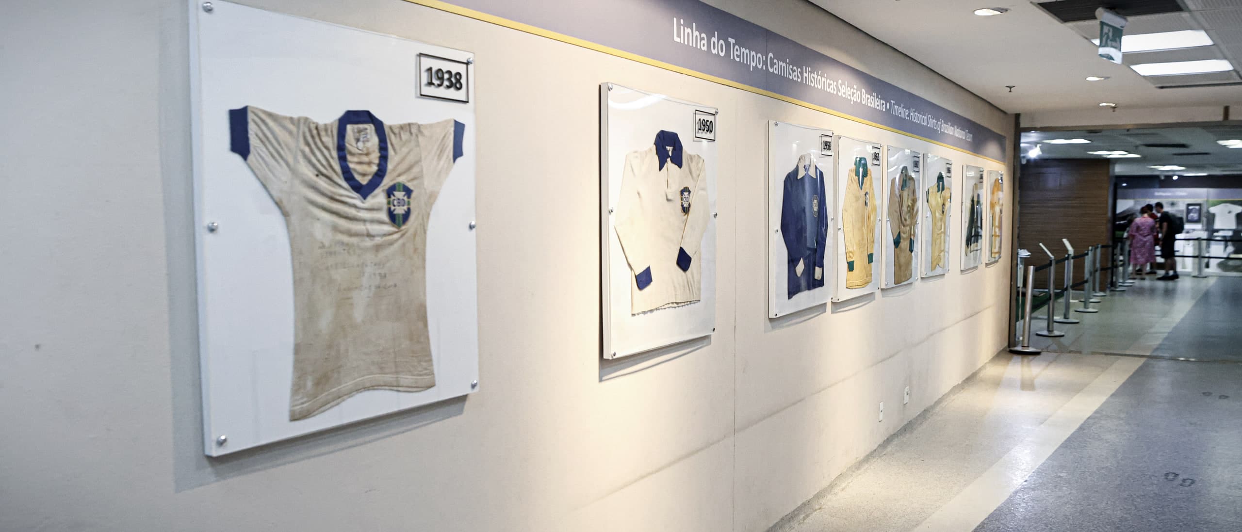 A Linha do Tempo das camisas da Seleção no museu do Maracanã: camisas emolduradas na parede, a de 1938 em primeiro plano, e o corredor de piso plano seguindo até o fundo.