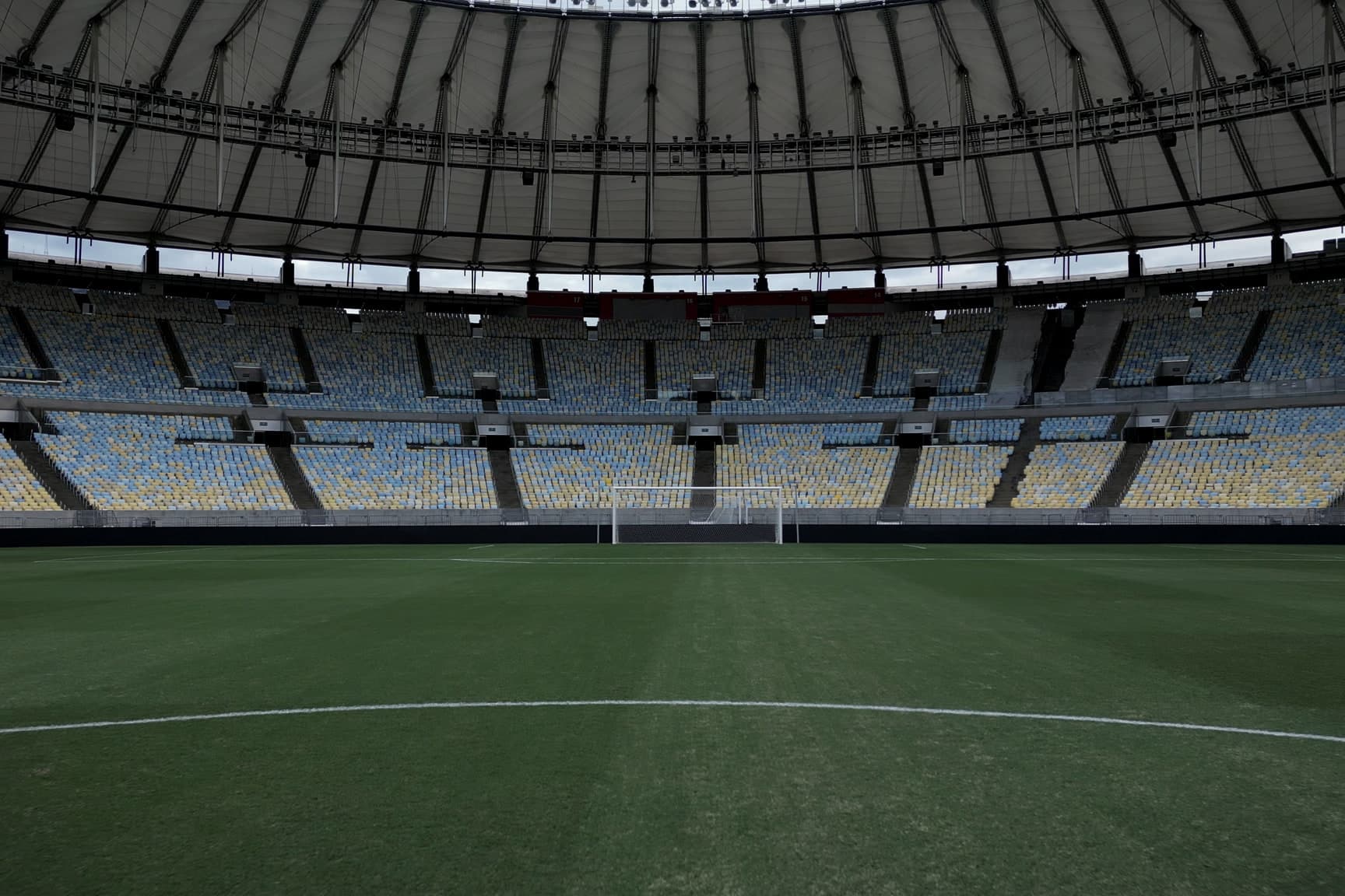 O gramado do Maracanã visto rente à grama, do círculo central em direção a um dos gols, com as arquibancadas vazias sob a cobertura.