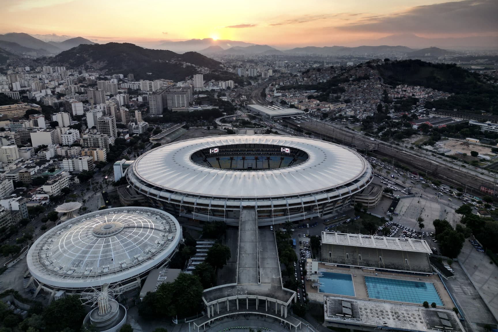 O Maracanã visto do alto ao pôr do sol, com o Maracanãzinho e o parque aquático à frente e a cidade até a serra ao fundo.