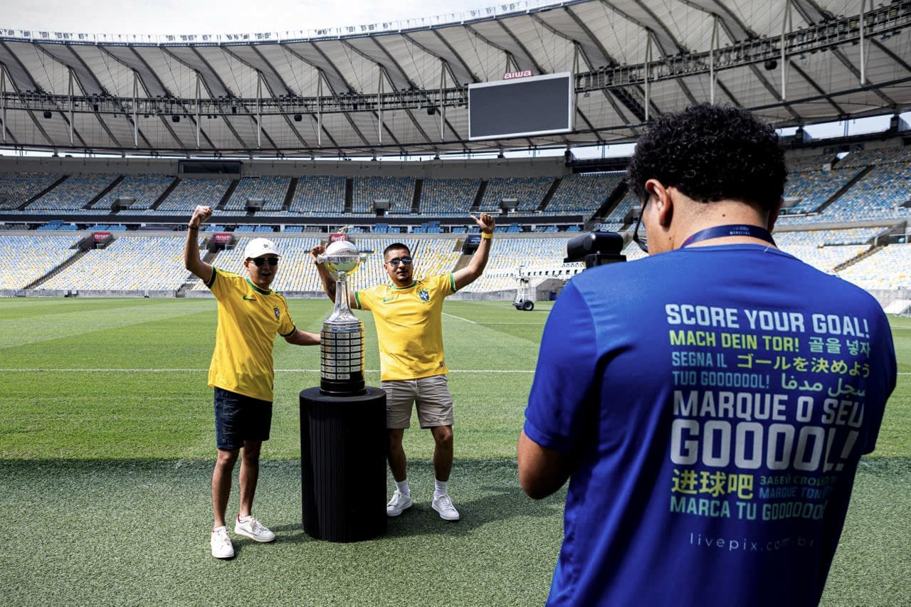 No gramado do Maracanã, um membro da equipe registra dois visitantes de camisa amarela posando ao lado da taça da Libertadores.
