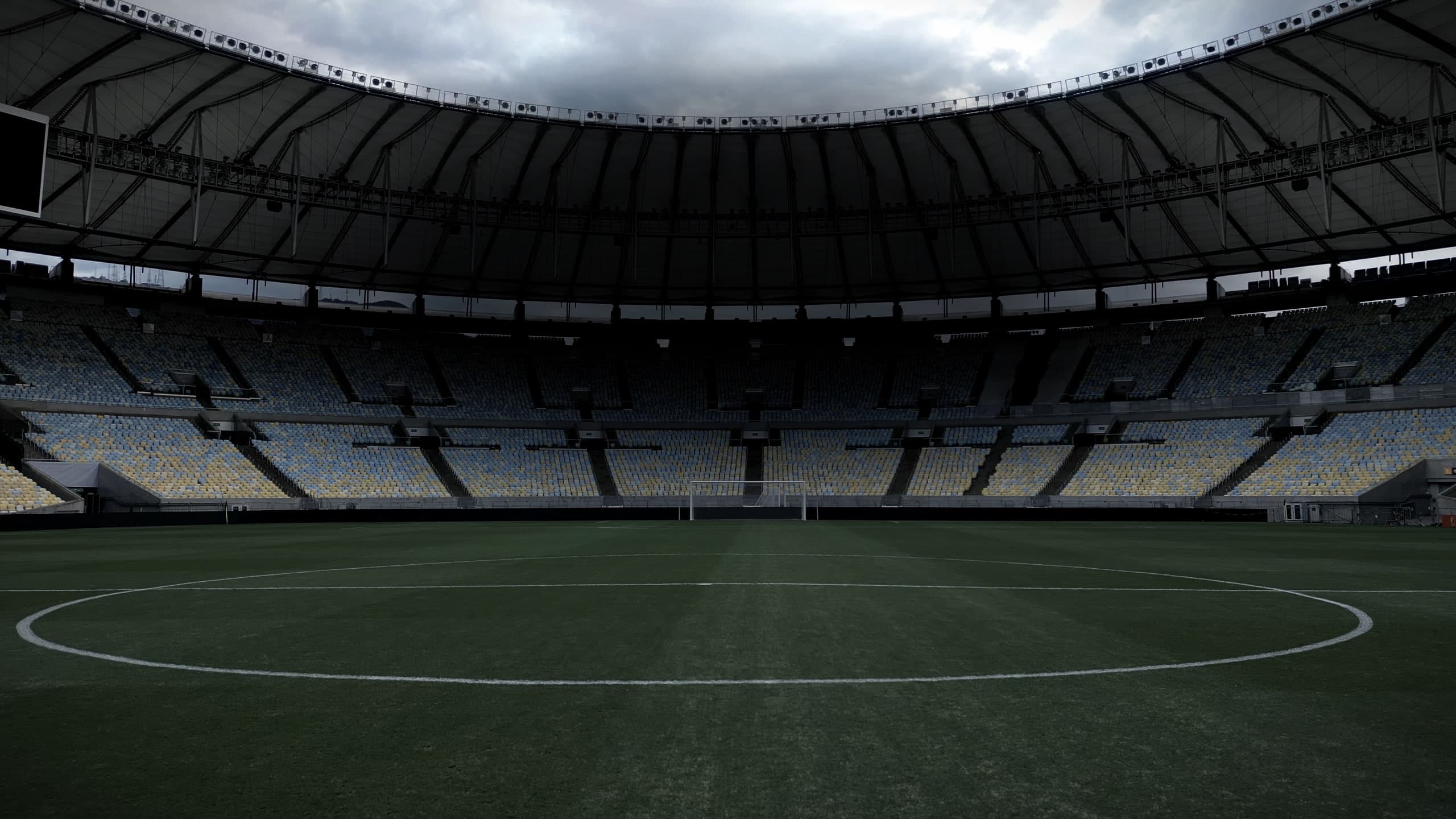 O gramado do Maracanã visto rente à grama, com o círculo central em primeiro plano e o anel inteiro de arquibancadas azuis e amarelas sob a cobertura, vazio.